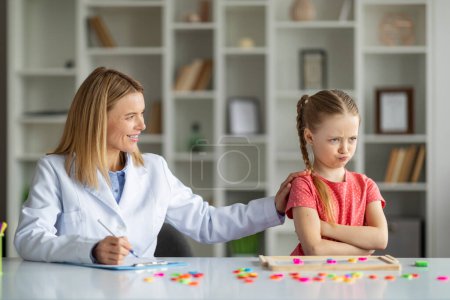 Photo for Behavior Therapy For Kids. Smiling Psychotherapist Woman Working With Grumpy Little Girl At Meeting In Office, Offended Female Child Standing With Folded Arms, Ignoring Therapist Lady - Royalty Free Image
