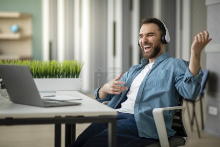 Téléchargez les photos : Joyeux jeune homme d'affaires qui s'amuse sur le lieu de travail au bureau, entrepreneur masculin positif écoutant de la musique dans des écouteurs sans fil et jouant de la guitare virtuelle, profitant d'une pause au travail, espace libre - en image libre de droit