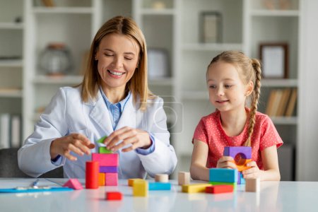 Photo for Child development specialist lady and cute little girl playing colorful wooden bricks together during therapy session in office, happy female kid enjoying development activities, closeup - Royalty Free Image