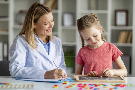 Photo for Smiling Little Girl Writting Letters On Mini Chalkboard During Meeting With Speech Therapist Lady At Office, Child Development Specialist Testing Female Child And Taking Notes, Closeup - Royalty Free Image