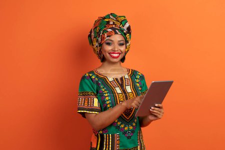 Téléchargez les photos : Joyeux joyeux millénaire femme noire en chemise africaine à l'aide d'une tablette numérique et souriant, isolé sur fond de studio orange. Assistant en ligne, chatbot, intelligence artificielle - en image libre de droit
