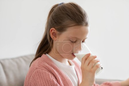 Photo for Caucasian teen girl pupil drinking glass of milk in living room interior, close up, free space. Health care, proper nutrition, diet, calcium for children at home, ad and offer - Royalty Free Image