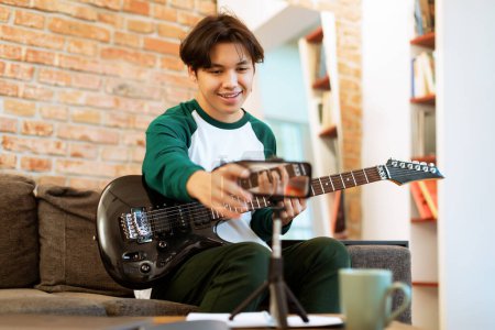 Photo for Internet Music Influencer. Chinese Teen Boy Records Guitar Session On Phone, Sharing Art And His Talent Online In Social Media, Sitting On Sofa At Home. Musical Blogging Concept. Selective Focus - Royalty Free Image