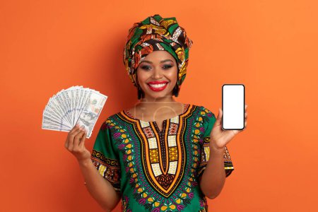 Téléchargez les photos : Cashback. Joyeux excité jolie jeune femme noire portant un costume africain national et un maquillage lumineux montrant smartphone avec écran blanc et dollars en espèces sur fond orange, maquette - en image libre de droit