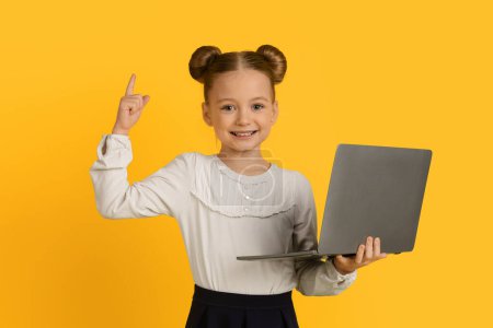 Téléchargez les photos : Concept d'idée. Petite écolière mignonne tenant un ordinateur portable et levant le doigt vers le haut, Happy Preteen Female Kid bénéficiant d'un site Web éducatif, Utilisation de l'ordinateur pour l'enseignement à distance, Debout sur fond jaune - en image libre de droit