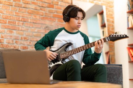Téléchargez les photos : Éducation musicale. Asiatique adolescent gars ayant des leçons de guitare en ligne, posant avec guitare et ordinateur portable, pratique assis à la maison. Tourné de sérieux étudiant de l'université de musique avec son instrument - en image libre de droit