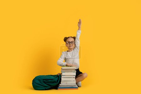 Téléchargez les photos : Petite écolière nerveuse mignonne assise avec une pile de livres et levant la main, une adolescente souriante répondant à la question sur la leçon, appréciant les connaissances et l'étude, posant sur un fond jaune - en image libre de droit