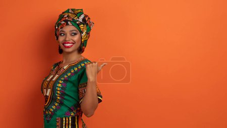 Photo for Positive happy beautiful young black woman wearing traditional arican clothing and bright makeup pointing with thumb at copy space for advertisement, isolated on orange background, web-banner - Royalty Free Image