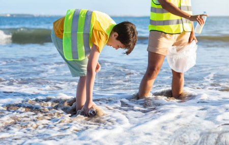 Foto de Triste adolescente caucásico voluntario en chaleco con maestro recoger basura y botellas de plástico de agua en la playa. Reciclaje, cuidado del medio ambiente, trabajo ecológico y ayuda a la naturaleza - Imagen libre de derechos