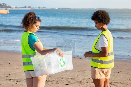 Foto de Triste adolescente grave diferente chica y señora voluntarios en chalecos recoger basura y botellas en caja en la playa. Trabajo ecológico, reciclaje de plástico, limpieza, ayuda a la naturaleza y el trabajo en equipo, cuidado del medio ambiente - Imagen libre de derechos