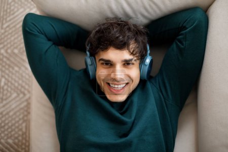 Téléchargez les photos : Portrait de jeune homme arabe heureux avec écouteurs sans fil bénéficie de musique préférée, confort et temps libre seul dans la chambre couché sur le canapé à la maison, regardant la caméra, prise de vue grand angle - en image libre de droit
