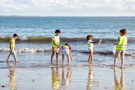 Téléchargez les photos : Occupé adolescent sérieux différents enfants bénévoles dans des gilets collectent les ordures et les bouteilles de l'océan sur la plage. Travail d'équipe, protection de l'environnement, éco-travail, recyclage du plastique, grotte naturelle - en image libre de droit