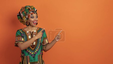 Foto de Emocionado alegre alegre hermosa mujer negra milenaria en traje africano tradicional apuntando al espacio de copia y mueca aislado en fondo de estudio naranja, mostrando buen trato, panorama - Imagen libre de derechos