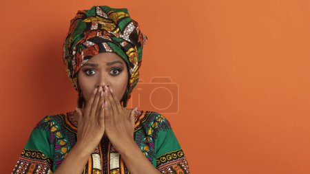 Foto de Mujer negra atractiva emocional horrorizada vistiendo maquillaje brillante y disfraz africano cubriéndose la boca, mostrando shock o asombro, panorama, espacio para copiar, fondo naranja - Imagen libre de derechos