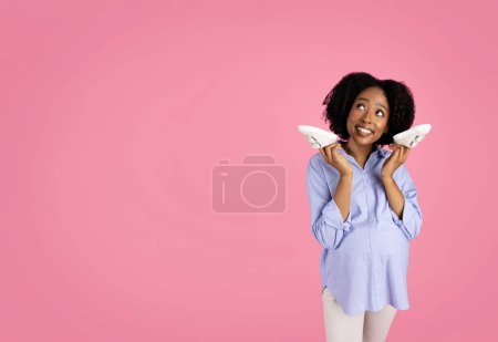 Photo for Smiling millennial african american curly pregnant woman in casual hold baby shoes, enjoy expect baby, future motherhood, shopping, look at free space, isolated on pink studio background - Royalty Free Image