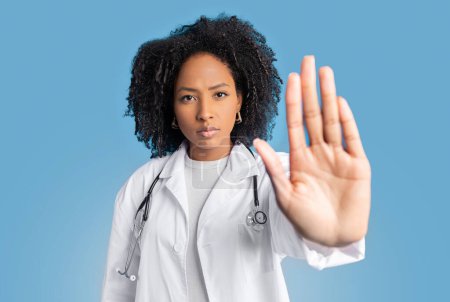 Téléchargez les photos : Sérieux confiant jeune afro-américaine bouclée médecin thérapeute en manteau blanc ne stop signe avec la main, isolé sur fond bleu, studio. Fin des problèmes, lutte contre les maladies, soins de santé - en image libre de droit