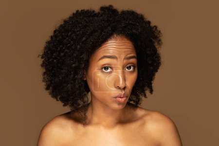 Foto de Mujer negra milenaria hermosa semidesnuda con expresión facial inocente posando aislada sobre fondo de estudio marrón, frente arrugada y mostrando labios de pato. Antienvejecimiento precoz belleza - Imagen libre de derechos
