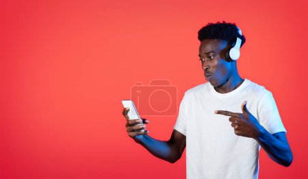Téléchargez les photos : Application musicale mobile. Incroyable excité cool jeune homme noir portant t-shirt blanc à l'aide d'écouteurs sans fil et pointant vers le smartphone dans sa main sur fond rouge dans la lumière au néon, panorama, espace de copie - en image libre de droit
