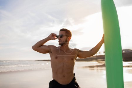 Foto de Muscular joven surfista chico posando en la playa al atardecer, guapo millennial hombre en elegante gafas de sol y traje de neopreno celebración de tabla de surf y mirando al lado, disfrutando de verano surf, Copiar espacio - Imagen libre de derechos