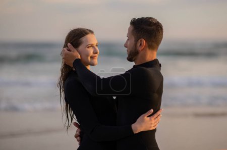 Foto de Romance junto a la playa. Retrato de una pareja romántica de jóvenes surfistas abrazándose en la playa, un hombre y una mujer felices y cariñosos en trajes de neopreno abrazándose y mirándose, disfrutando del surf juntos, espacio libre - Imagen libre de derechos