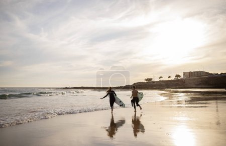 Téléchargez les photos : Wave Runners. Jeune couple avec planches de surf allant dans l'océan, Millennial Homme et femme en combinaison Surfer sur le coucher du soleil, Courir à haute vagues, S'amuser sur la plage ensemble, Copier l'espace - en image libre de droit