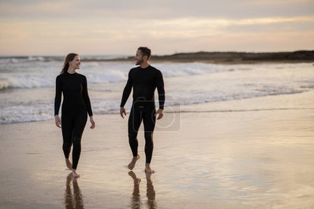 Foto de Feliz joven hombre y mujer en trajes de neopreno caminando por la playa al atardecer, pareja milenaria romántica relajándose a la orilla del mar, saliendo del agua después de surfear juntos, de cuerpo entero, espacio para copiar - Imagen libre de derechos