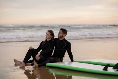 Photo for Happy young couple chilling out on the beach after surfing, millennial surfers man and woman wearing wetsuits sitting on sand near their boards, romantic lovers looking at sunset, copy space - Royalty Free Image