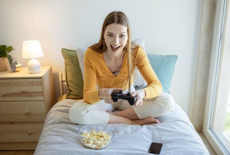 Téléchargez les photos : Jeux vidéo Divertissement. Jeune fille blonde excitée jouant au jeu vidéo en ligne Holding Game Controller et s'amuser le week-end, assis sur le lit à la maison. Concept de technologie et loisirs numériques - en image libre de droit