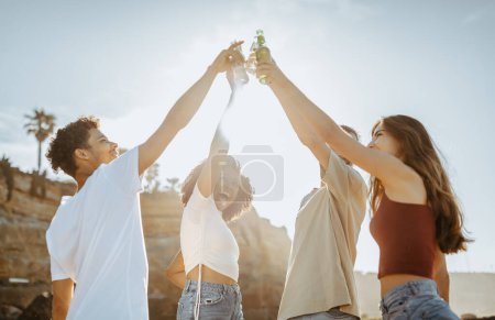 Photo for Cheerful millennial international people students cheers with bottles, enjoy summer holidays, picnic on beach, sun flare. Freedom lifestyle, free time at weekend, party together and fun - Royalty Free Image
