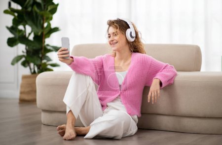 Photo for Relaxed young woman listening music on smartphone and wireless headphones while relaxing on floor at home, smiling lady enjoying favorite songs, downoading playlist, resting in living room - Royalty Free Image