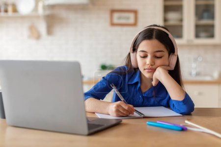 Photo for Boring Online Education. Bored Schooler Girl Learning At Laptop And Taking Notes Sitting With Earphones At Desk At Home. Tired School Kid Doing Difficult Homework Indoors - Royalty Free Image
