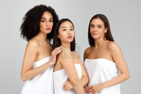 Téléchargez les photos : Beauté féminine multiraciale naturelle. Portrait de trois dames à la peau douce et parfaite, posant enveloppé dans une serviette, regardant la caméra sur fond gris, prise de vue en studio, bannière - en image libre de droit