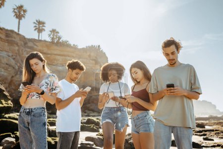 Foto de Triste millennial negro, asiático, árabe y europeos estudiantes en las vacaciones de verano, charlando en los teléfonos inteligentes en la playa. Blog, aplicación de redes sociales, problemas en fin de semana, mapa en línea en viaje - Imagen libre de derechos