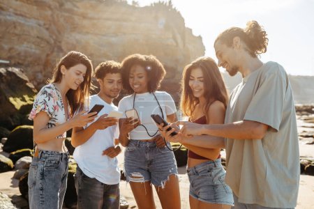 Photo for Smiling millennial multiracial people student enjoy summer holidays, chatting in social networks on phones on beach. Game, website recommendation, blog, app and modern lifestyle, weekend together - Royalty Free Image