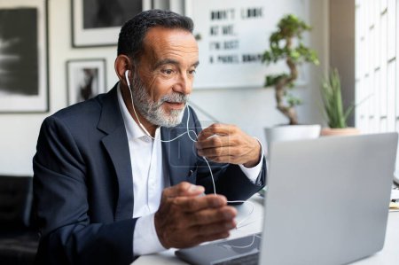 Photo for Cheerful mature european man in suit and headphones working on laptop in modern office interior. Meeting, business remotely, device for chat, communication with client, support and financial advice - Royalty Free Image