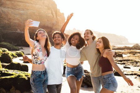 Foto de Feliz millennial multiétnicos estudiantes disfrutan de vacaciones de verano, divertirse, tomar selfie en el teléfono en la playa. Blog de viajes, foto para redes sociales, aplicación, estilo de vida activo juntos - Imagen libre de derechos