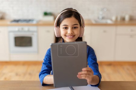 Foto de Concepto de Educación Web. Chica alegre de la escuela sosteniendo tableta digital utilizando el sitio web educativo, haciendo la tarea con auriculares, sentado en el escritorio en casa. Retrato de niño feliz con almohadilla - Imagen libre de derechos