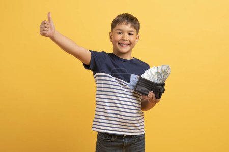 Photo for Happy rich cute boy schooler in casual outfit holding black leather wallet full of cash dollar banknotes and showing thumb up, isolated on yellow studio background. Kids and financial literacy - Royalty Free Image