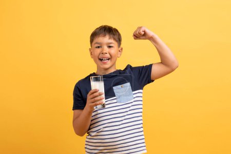 Photo for Adorable happy caucasian school aged boy with glass of milk posing on yellow studio background, showing strong muscles, demosntrating biceps. Kids nutrition, healthy diet for children - Royalty Free Image