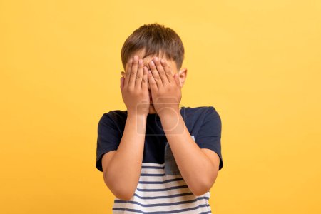 Photo for Shy boy feeling ashamed because of his bad behavior, crying, not showing his tears. Isolated shot of unrecognizable kid with hands on face, afraid of scary movie, yellow background. Body language - Royalty Free Image