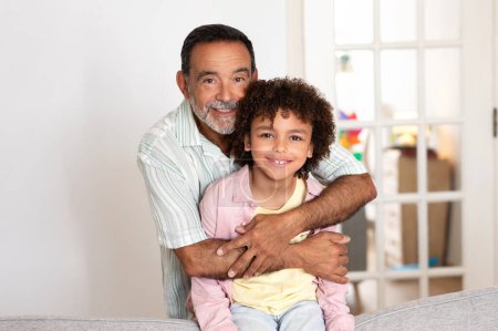 Foto de Abuelo y nietos retrato. Feliz abuelo latino abrazando a su nieto posando juntos y sonriendo en la cámara de pie en la sala de estar moderna en casa. Bono familiar, Generaciones - Imagen libre de derechos