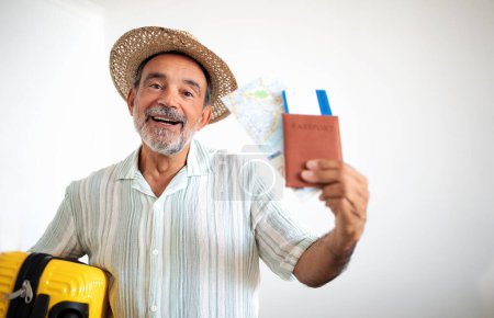 Foto de Viajar en cualquier época. Hombre adulto hispano jubiloso con sombrero de verano y maleta amarilla mostrando pasaporte con boletos de avión de tarjeta de embarque, sonriendo ante la cámara posando contra fondo gris de la pared - Imagen libre de derechos