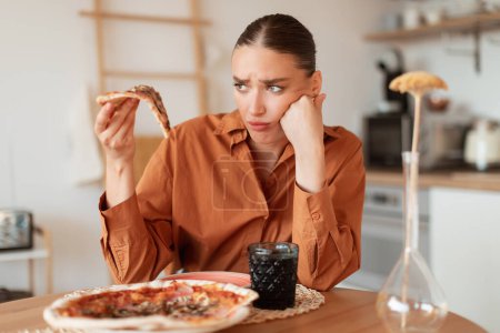 Téléchargez les photos : Femme bouleversée regardant un délicieux morceau de pizza à haute teneur en calories, tenant une tranche à la main. Problèmes alimentaires et digestifs ainsi que des aliments gâtés et insipides - en image libre de droit