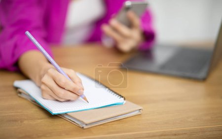 Photo for Planning Concept. Closeup Of Female Writing In Notepad And Holding Smartphone While Sitting At Desk In Office, Unrecognizable Woman TaKing Notes To Notebook, Making To-Do List, Cropped - Royalty Free Image