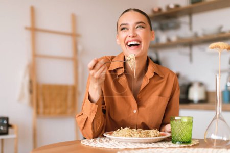 Photo for Kitchen happenings. Excited woman eating tasty homemade pasta, enjoying delicious lunch and laughing, sitting in cozy kitchen interior, free space - Royalty Free Image