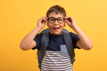 Photo for Closeup of emotional excited happy kid scooler boy with backpack behind his back adjusting eyewear and looking at camera, isolated on yellow studio background. School, kids education - Royalty Free Image
