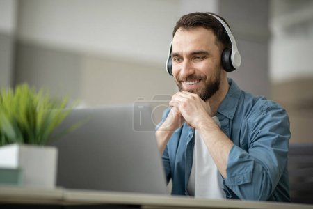 Téléchargez les photos : Un jeune homme souriant dans un casque sans fil regardant un webinaire sur un ordinateur portable, un homme heureux et millénaire bénéficiant d'une éducation en ligne et d'un enseignement à distance, assis au bureau et regardant l'écran d'ordinateur, de l'espace libre - en image libre de droit