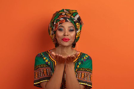 Photo for Flirtatious funny beautiful young african american lady wearing national costume and bright makeup holding hands next to lips, sending fly kiss at camera isolated on orange background - Royalty Free Image
