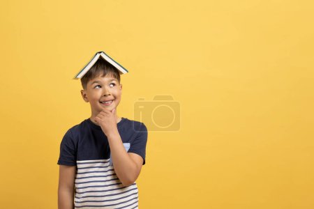 Foto de Feliz lindo guapo chico de edad escuela caucásica en traje casual con libro en la cabeza tocándose la barbilla y mirando el espacio de copia, aislado en el fondo amarillo. Literatura infantil - Imagen libre de derechos