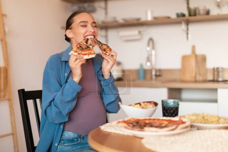Téléchargez les photos : Jeune femme enceinte excitée dégustant une pizza, mordant deux tranches savoureuses, assise à table à l'intérieur de la cuisine, espace de copie. Amateurs de malbouffe mangeant de la pizza italienne. Alimentation malsaine tricher repas - en image libre de droit